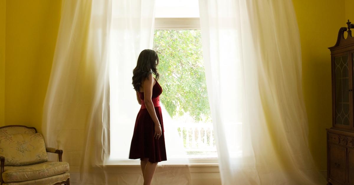 woman stands looking outside at big open window with billowing curtains